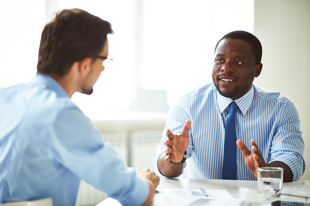 A man in an office suit and tie speaking to another man wearing glasses.