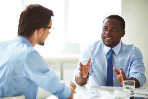 A man in an office suit and tie speaking to another man wearing glasses.