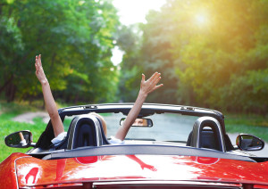 A woman driving a red car raising her arms as she drives through a forest.