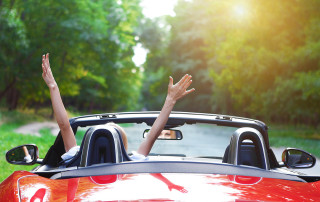 A woman driving a red car raising her arms as she drives through a forest.
