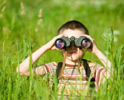 A little boy in a striped shirt hiding amongst tall grass and looking through binoculars.