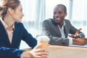A man and a woman sitting together by a conference table talking and smiling.