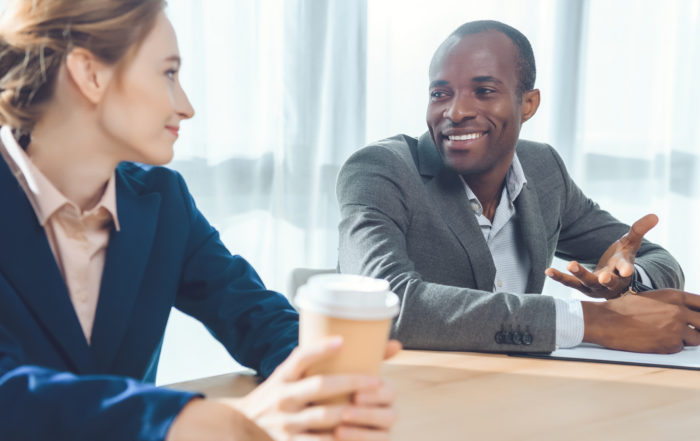 A man and a woman sitting together by a conference table talking and smiling.